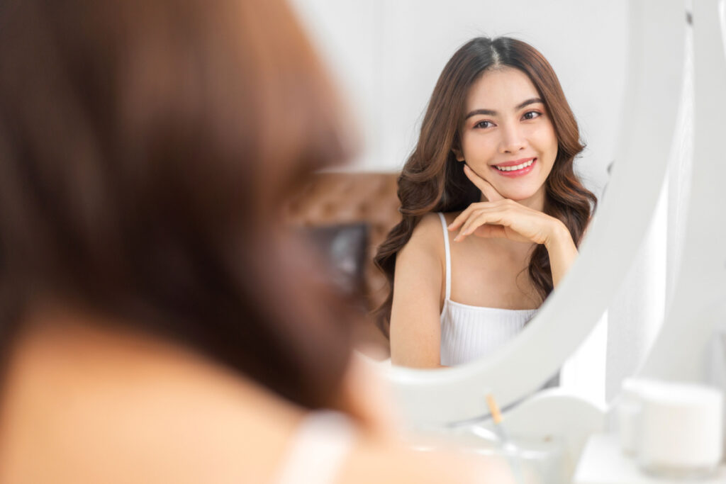 Woman admiring her reflection, considering the costs of buccal fat removal surgery.