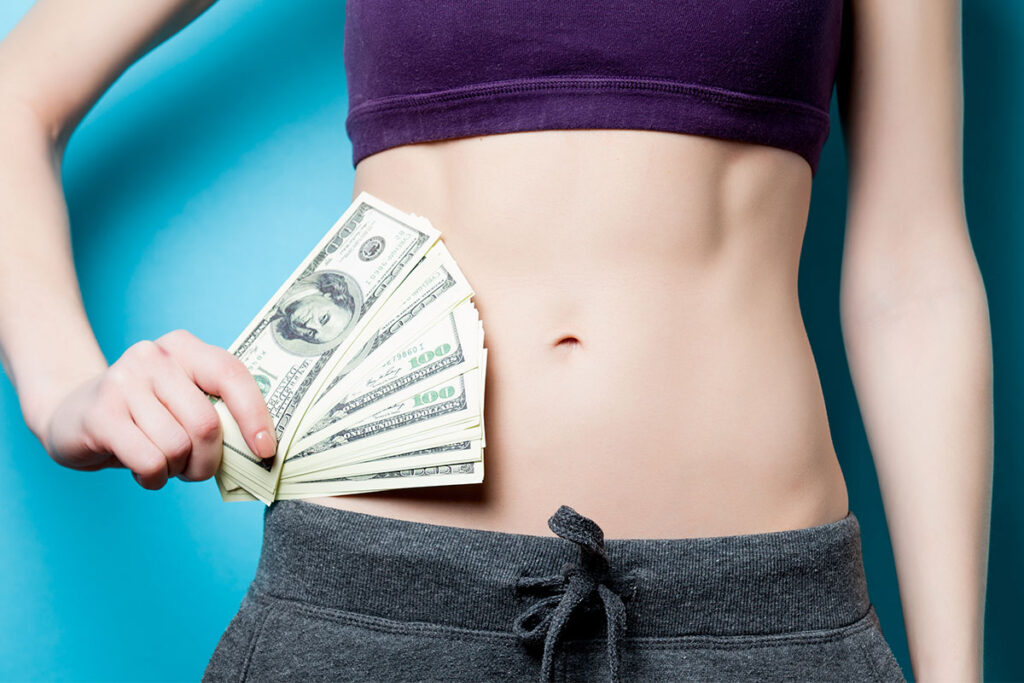 Frontal view of a woman's abdomen while she holds dollar bills in relation to financing options after extra skin removal surgery or body lift surgery.